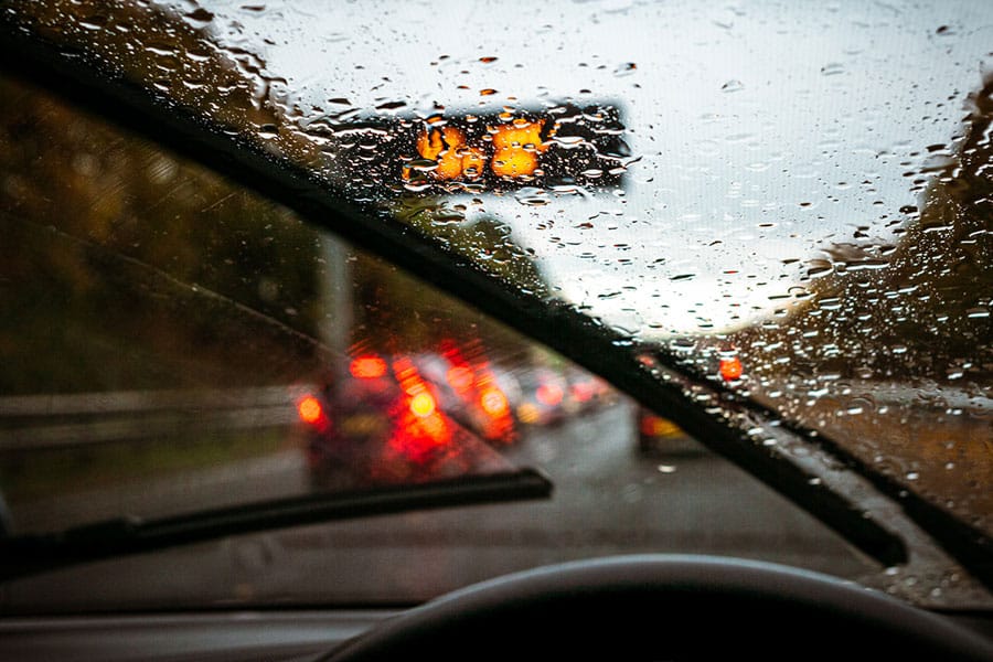 View from inside a car during rain, with windshield wipers in motion. Traffic with red brake lights is visible ahead, and a blurry illuminated sign can be seen through the wet windshield—reminding drivers of common car wash myths and the importance of proper car cleaning to maintain clear visibility.