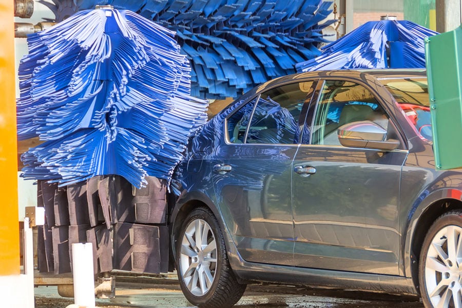 A dark-colored car is being washed in an automatic Drip Car Wash, surrounded by large blue spinning brushes and cared for by a dedicated team.