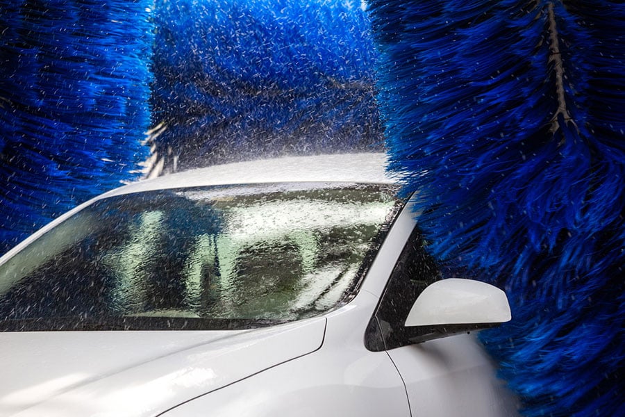 A white car is being washed in an automatic Drip Car Wash, surrounded by large spinning blue brushes with water spraying everywhere—a glimpse into the day in the life of their dedicated team ensuring every vehicle shines.