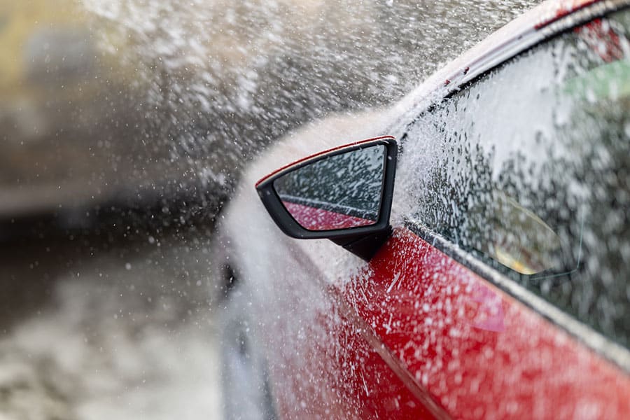 Close-up of a red car’s side mirror with water spraying over it, likely during a car wash at Drip Car Wash—just another day in the life for our dedicated team.