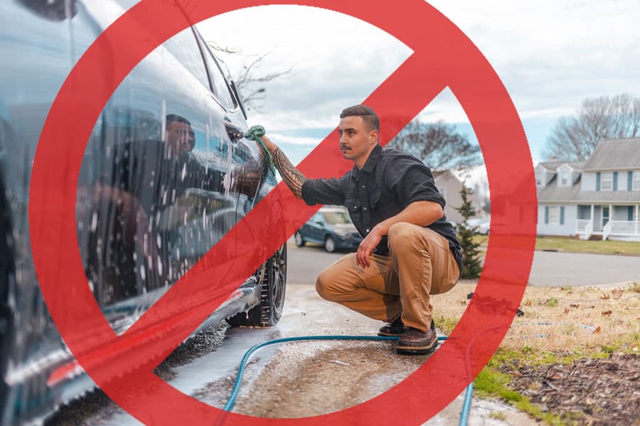 A man washing a car outdoors with soap and sponge, overlaid by a large red prohibition sign, indicating that washing cars in this manner is not allowed in Michigan, where Eco-Friendly Car Care practices—such as using a Drip Car Wash—are encouraged.