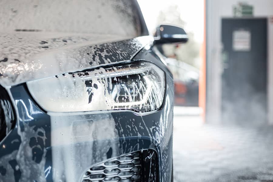 Close-up of a car’s front headlight covered in soap suds during a wash, highlighting effective car wash techniques, with a blurred background showing part of an open garage and another vehicle.