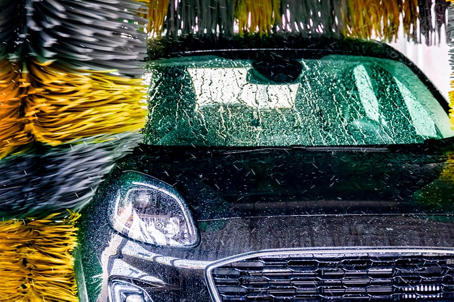 A black car is being washed in an automatic car wash, with large yellow and gray brushes cleaning the windshield and hood—an essential routine for many Michigan drivers dealing with the area's changing climate.