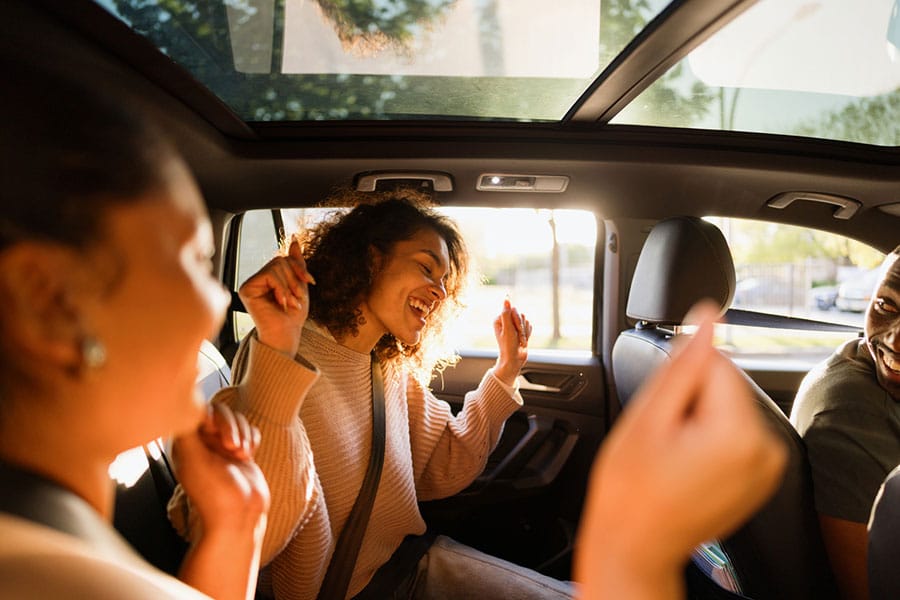 Three people enjoying music in a car, with one woman in the backseat smiling and dancing, sunlight streaming through the windows, and everyone appearing happy and relaxed—all while cruising through Michigan communities. Their shared joy reflects the spirit of supporting local businesses like Drip Car Wash.