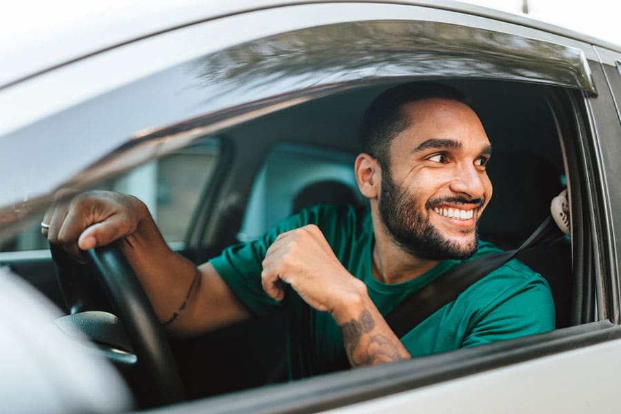 A smiling man in a green shirt sits in the driver’s seat of a car, looking out the open window and holding the steering wheel with one hand—showcasing the results of diligent Michigan car care.