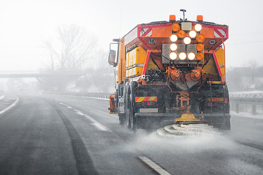 A large orange road salt spreader truck spreads salt on a snowy, icy highway during winter—a vital part of winter car care for Michigan drivers, with snow visible on the ground and trees in the background.