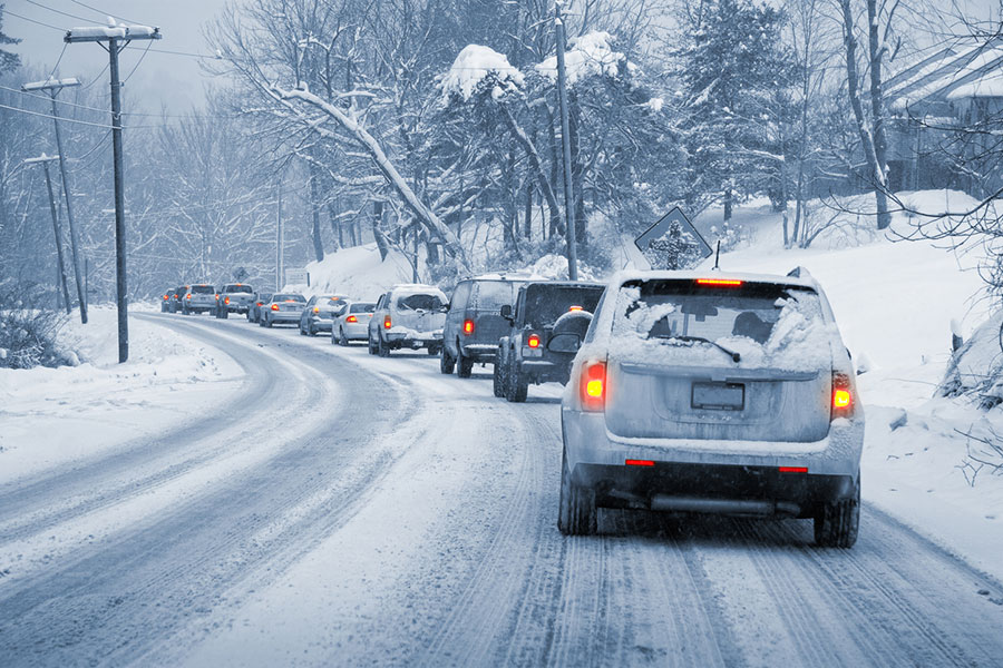 A line of cars drives slowly on a snow-covered, winding road through a snowy landscape with trees and houses. Snow is piled on vehicles and utility lines, indicating recent or ongoing snowfall—typical of Michigan winters, when it’s especially important to protect your car from road salt and grime.