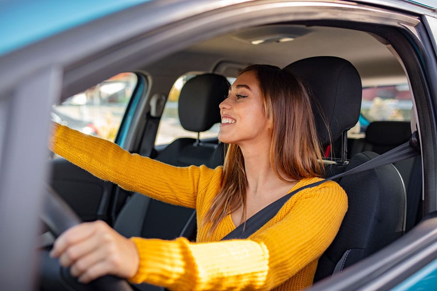 A young woman wearing a yellow sweater smiles while driving a car, looking out the window with her hands on the steering wheel, enjoying the shine of her vehicle thanks to regular car washing.